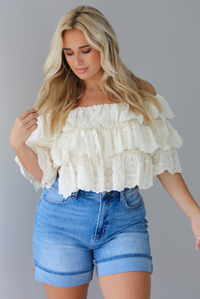 Woman wearing a cream off the shoulder, ruffled eyelet cropped top with medium wash denim shorts indoors against a white wall.