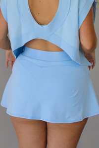 A woman wearing a light blue two-piece outfit consisting of a short-sleeve crop top with draped sleeves and a matching high-waisted mini skirt, standing indoors against a white wall. The photo shows a closeup of the back of the mini skirt.