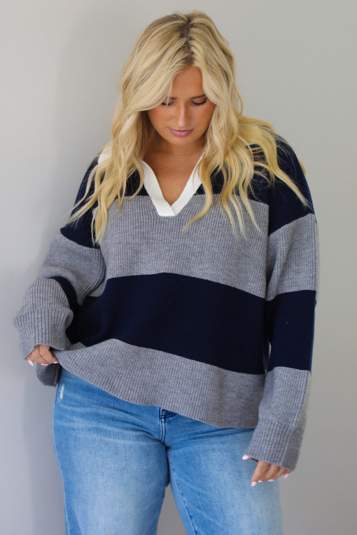 A woman wearing a navy and charcoal boyfriend style pullover top with a white collared detail, with distressed denim, standing indoors against a white wall.