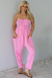 Woman wearing a pink strapless jumpsuit with a fitted bodice and slouchy pants indoors against a white wall with white marble flooring. 