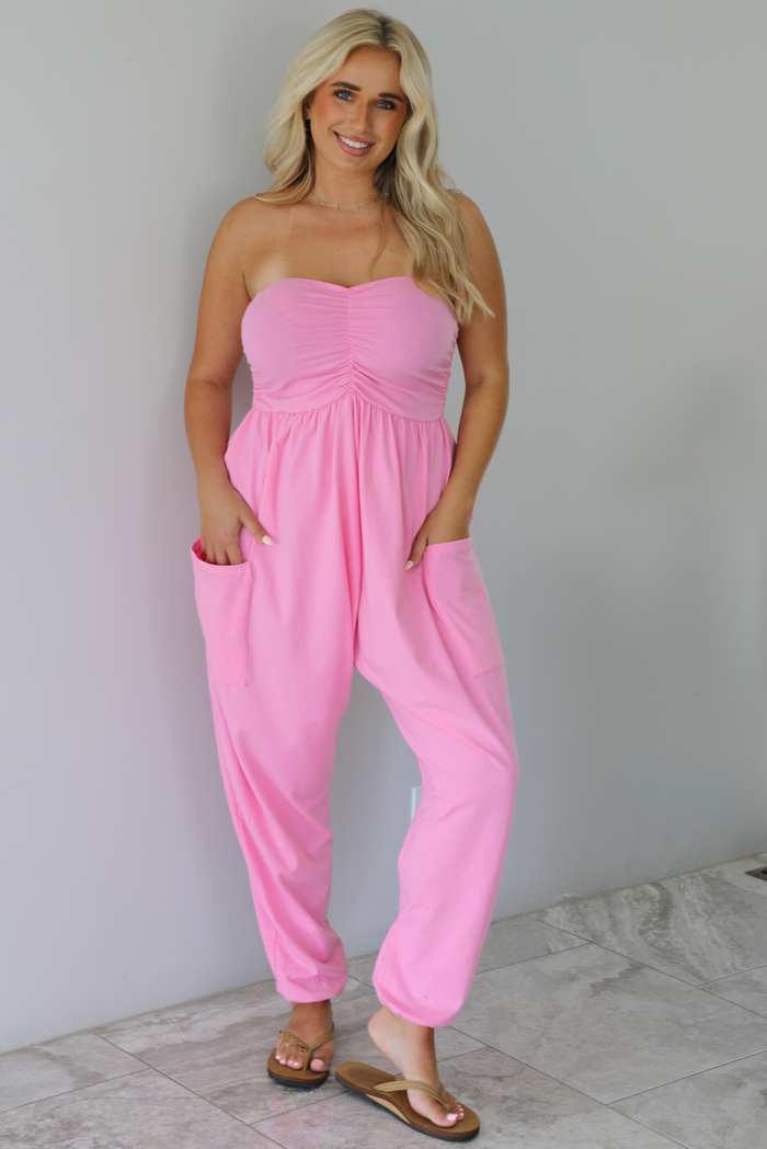 Woman wearing a pink strapless jumpsuit with a fitted bodice and slouchy pants indoors against a white wall with white marble flooring. 