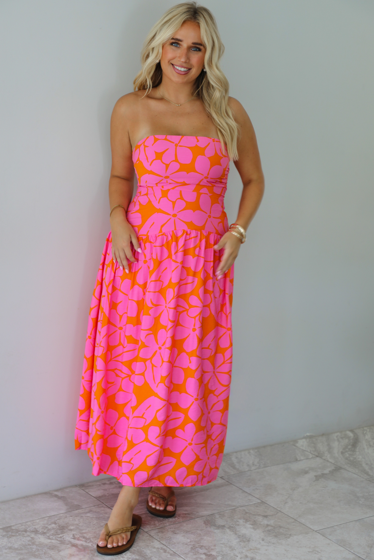 A woman modeling a strapless midi dress with a vibrant pink and orange floral pattern, standing indoors agains a white wall with white marble flooring. 