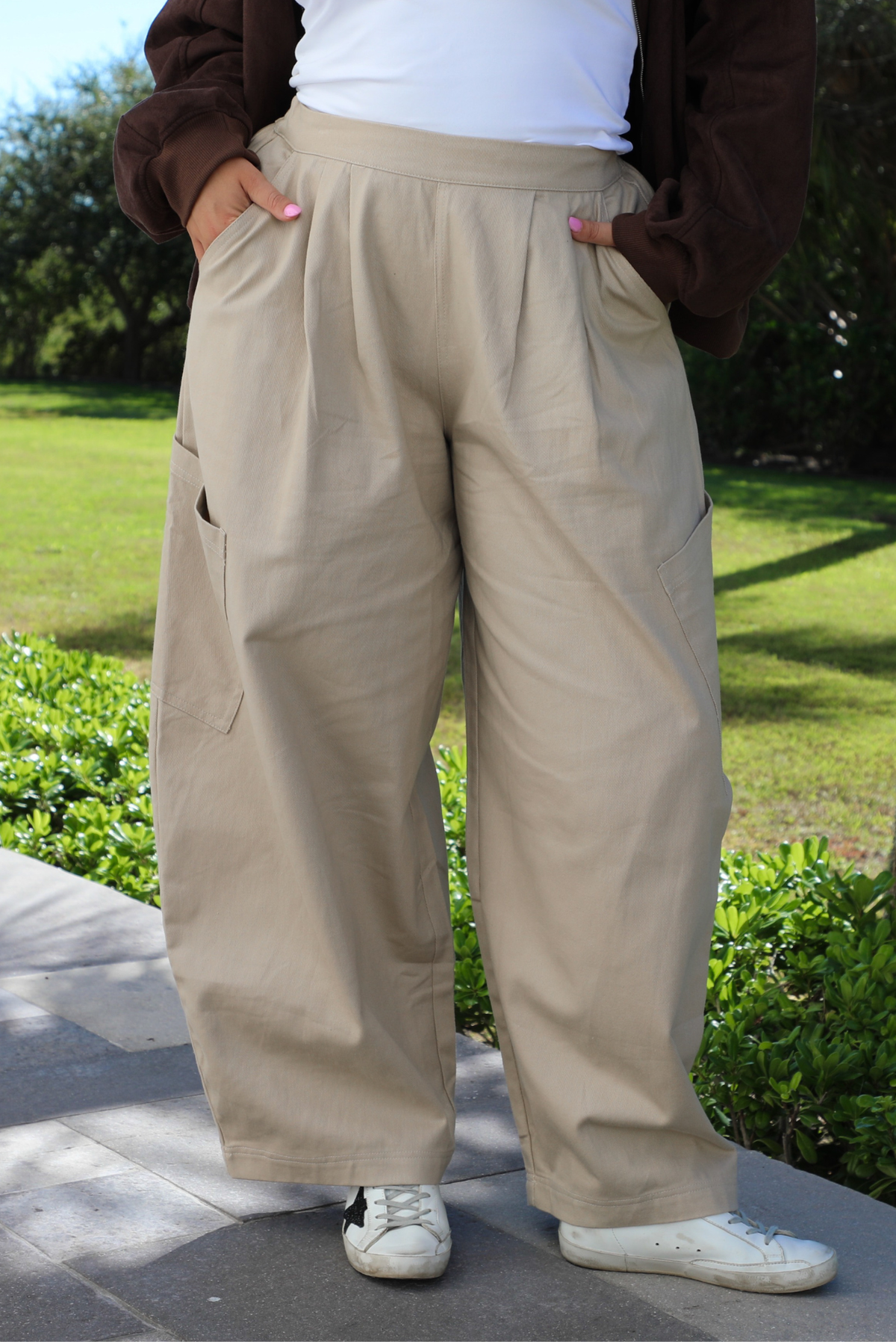 A woman shown from the waist down wearing a pair of sand baggy pants, standing outdoors on a stone path with greenery in the background. 