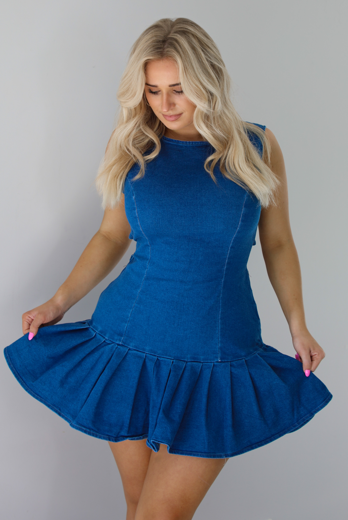 Woman wearing a dark blue denim dress that has a fitted bodice with a drop waist design thad leads to a pleated hem design. The photo shows a closeup of the dress. The photo is taken indoors against a white wall.