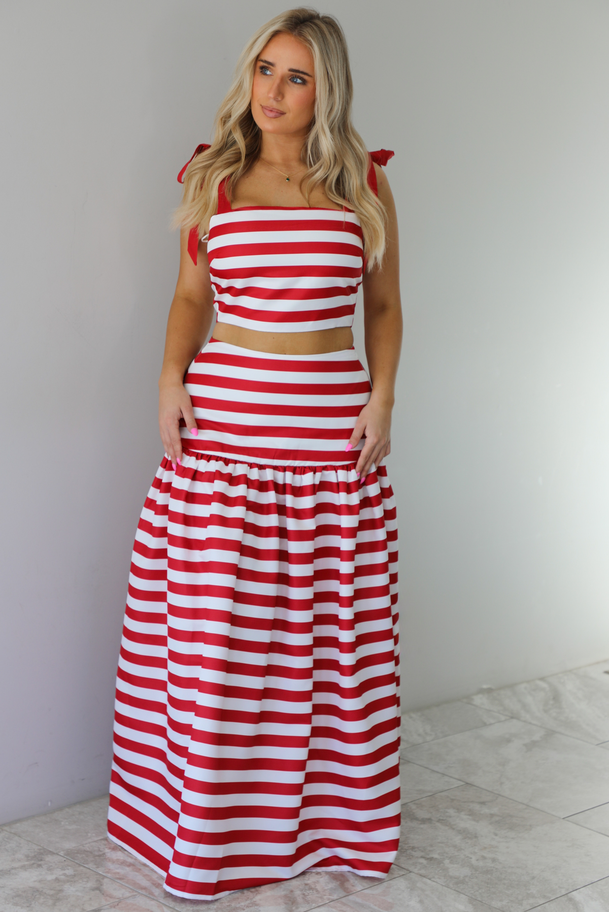 Woman wearing a red and white striped two piece set featuring a cropped top and maxi length, high waisted skirt. The photo is taken indoors against a white wall with white marble flooring. 