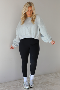Woman wearing a heather grey cropped sweatshirt with a black fitted jumpsuit underneath. The outfit is paired with high ankle socks and silver sneakers. The photo is taken indoors against a white wall with white marble flooring. 