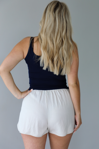 Woman wearing a navy ribbed tank  untucked with stone colored shorts indoors against a white wall. The photo shows the back of the top. 