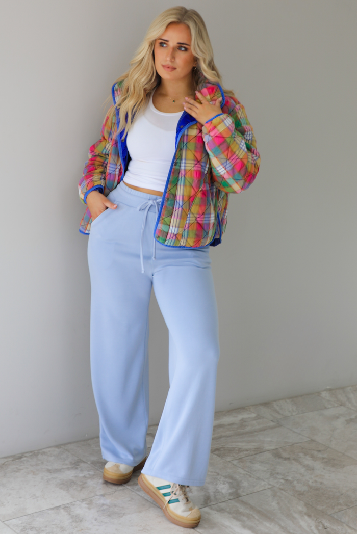 Woman wearing a multi-colored plaid, quilted style jacket paired with a white top underneath and high waisted blue lounge pants. The photo is taken indoors against a white wall with white marble flooring. 