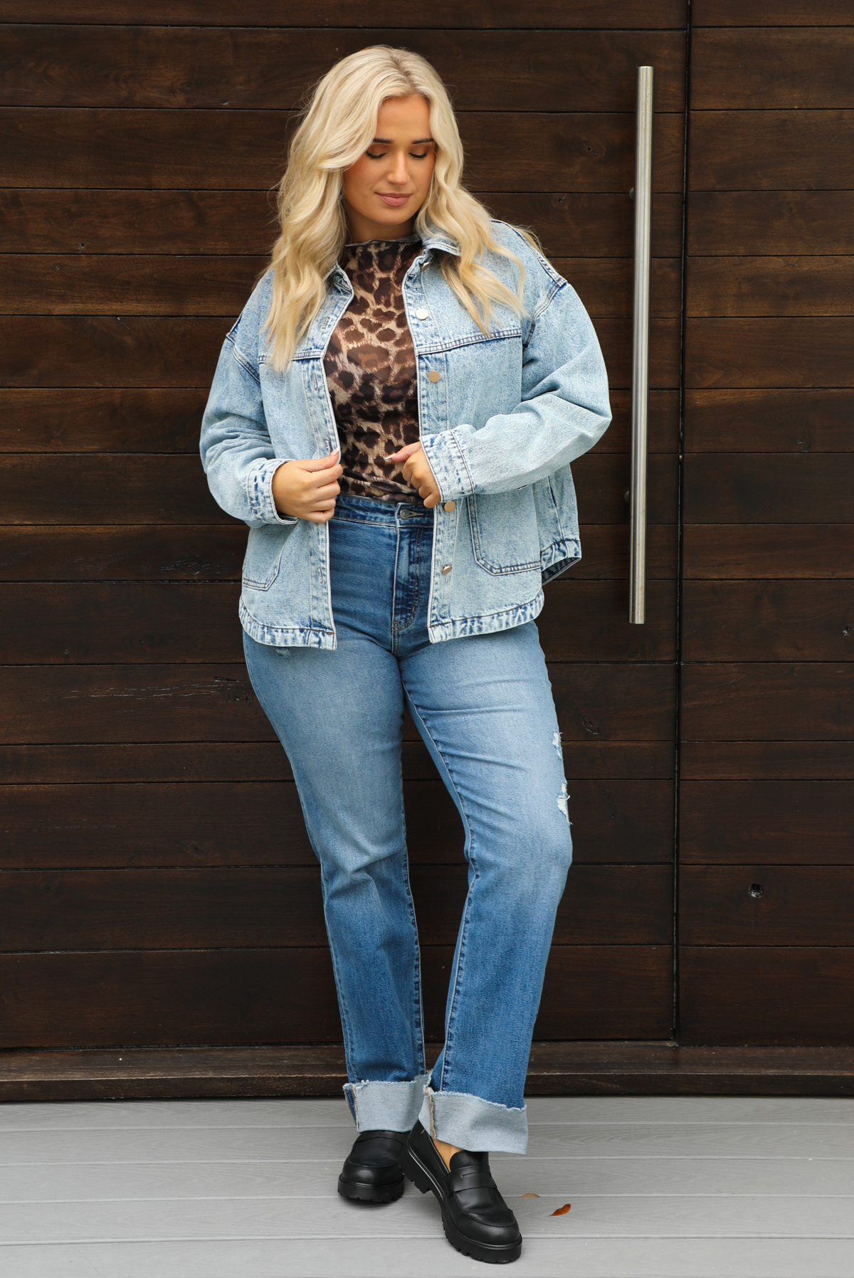 Woman model wearing an oversized, acid washed, light denim jacket, paired with a sheer mesh leopard print top, medium wash denim jeans, and black shoes. The photo is taken outdoors against a dark brown door. 