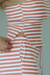 Woman wearing an orange, white, and green striped pattern set that has a strapless tube top and high waisted pants indoors against a white wall and shows a closeup of the side of the top, which has a split hem detail. 