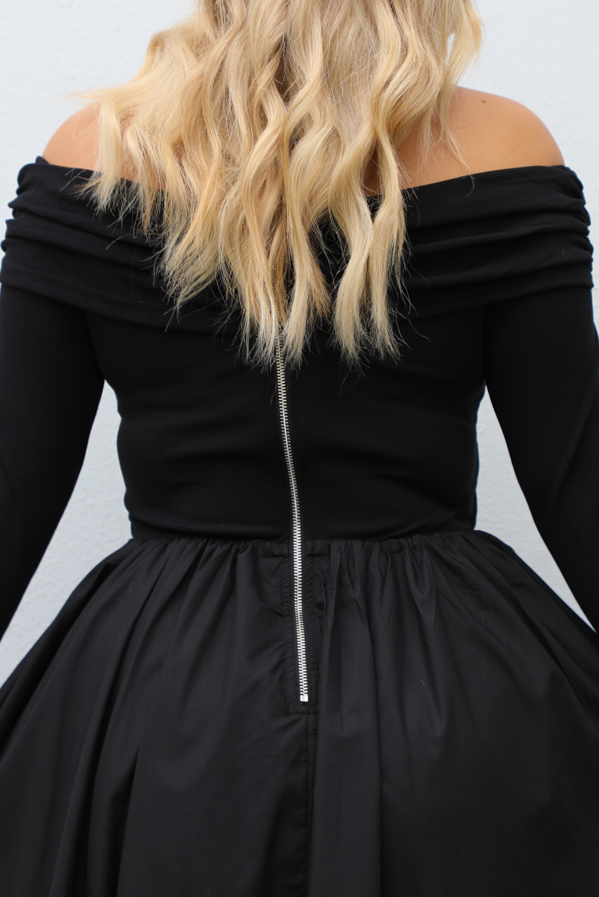 Woman wearing a black off the shoulder, long sleeved midi length dress with a pair of clear strapped nude heels. The photo shows a closeup of the back of the dress, which is similar to the front except it also has a zipper closure. The photo is taken outdoors against a white wall.