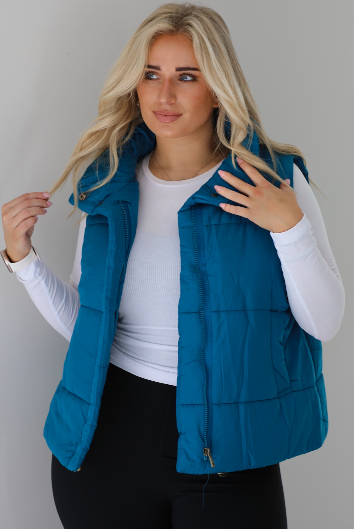 Woman wearing a teal puffer vest, white long-sleeve shirt, and black leggings indoors against a white wall. The puffer vest has a collared design, functional button closures at the neckline, an unzipped closure in the front, and functional pockets on either side.