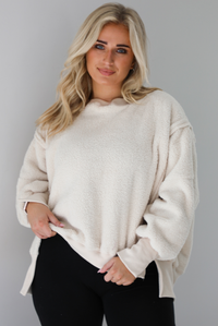 Woman wearing a sherpa sand oversized sweater with a fitted black jumpsuit underneath. The photo shows a closeup of the sweater and the raw seam details. The photo is taken indoors against a white wall with white marble flooring. 
