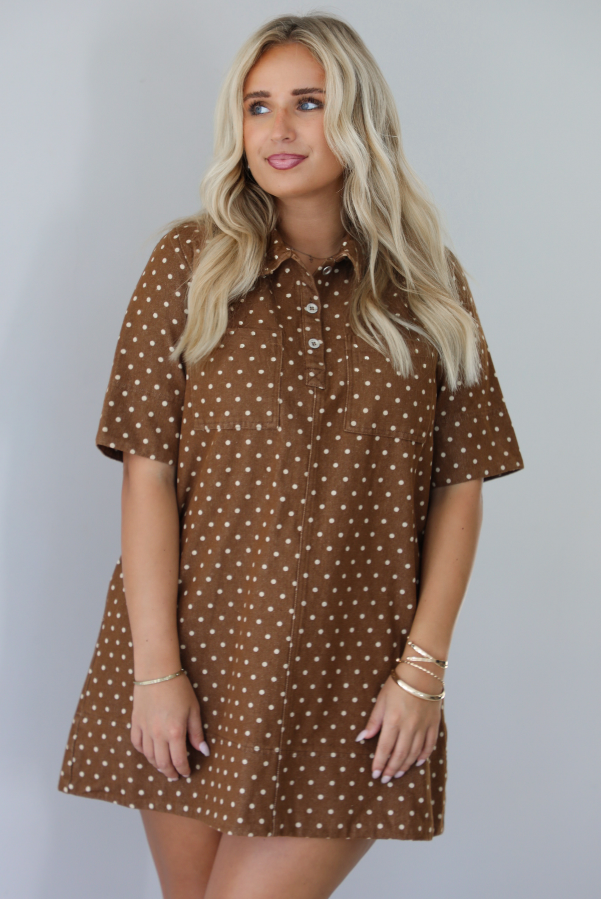 A woman modeling a brown short-sleeve mini dress with a cream polka dot pattern, standing indoors against a white wall.