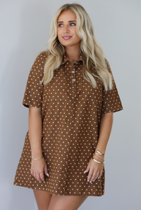 A woman modeling a brown short-sleeve mini dress with a cream polka dot pattern, standing indoors against a white wall.
