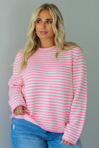 A woman wearing a pink and white striped sweater paired with light wash denim jeans, standing indoors against a white wall.