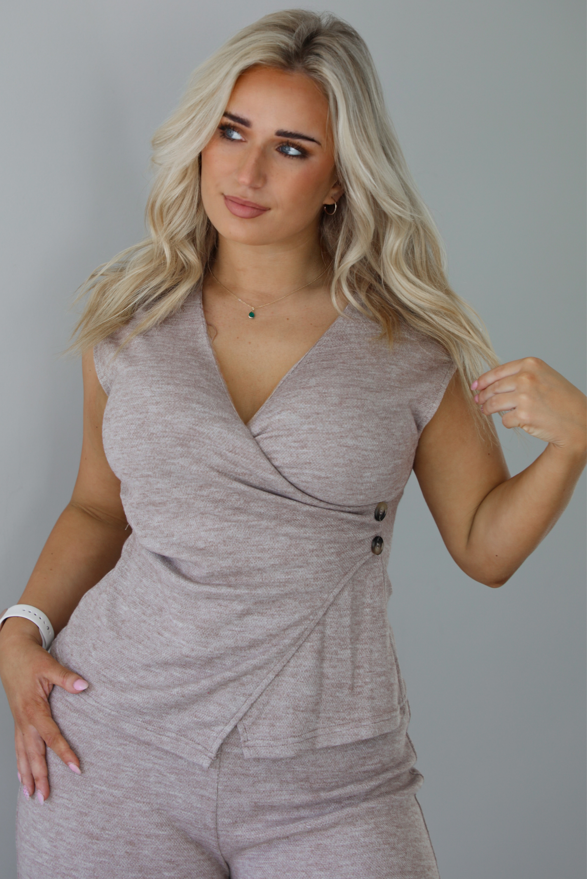 Woman wearing a taupe colored two piece set featuring a vest-appearance top with non-functional button closures on the left side and high waisted pants. The photo shows a closeup of the top. The photo is taken indoors against a white wall.