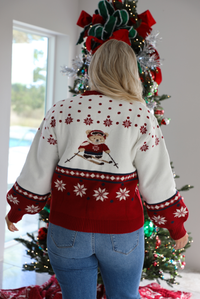 Back of a woman model wearing a red and white Christmas cardigan with teddy bears skiing with functional button closures in the front. The cardigan is styled with medium wash denim jeans that are cuffed at the hem. The photo is taken inside a modern home with a decorated Christmas tree in the back. 