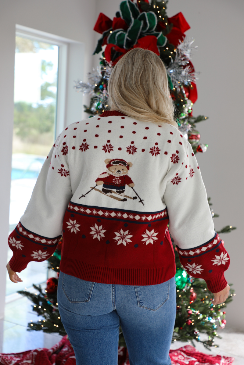Back of a woman model wearing a red and white Christmas cardigan with teddy bears skiing with functional button closures in the front. The cardigan is styled with medium wash denim jeans that are cuffed at the hem. The photo is taken inside a modern home with a decorated Christmas tree in the back. 