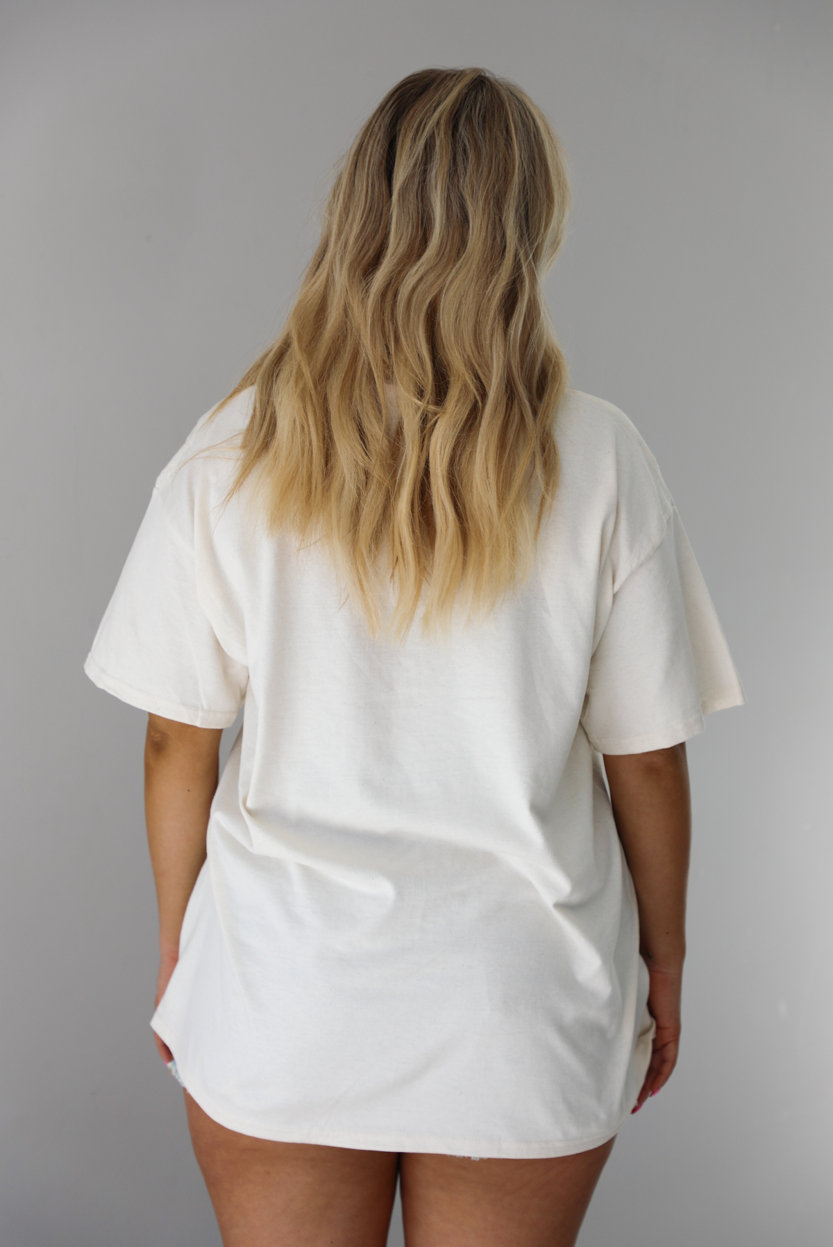 Woman wearing a white relaxed fit graphic tee that has various tickets on it with different fruits and flowers on them. The tee is untucked to show the full length and paired with distressed denim shorts. The photo shows the back of the tee, which is plain. The photo is taken indoors against a white wall.