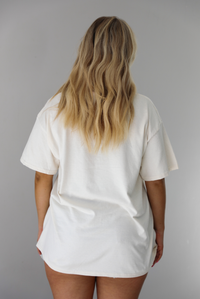 Woman wearing a white relaxed fit graphic tee that has various tickets on it with different fruits and flowers on them. The tee is untucked to show the full length and paired with distressed denim shorts. The photo shows the back of the tee, which is plain. The photo is taken indoors against a white wall.