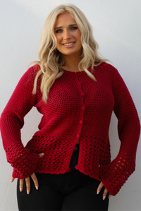 Close up of a woman model wearing a burgundy knit cardigan with button closures in the front and black denim jeans. The photo is taken outdoors against a white wall. 