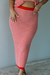 Woman wearing a red and white knit two piece set featuring a sleeveless top and high waisted midi length skirt indoors against a white wall with white marble flooring. The photo shows a closeup of the front of the skirt. 