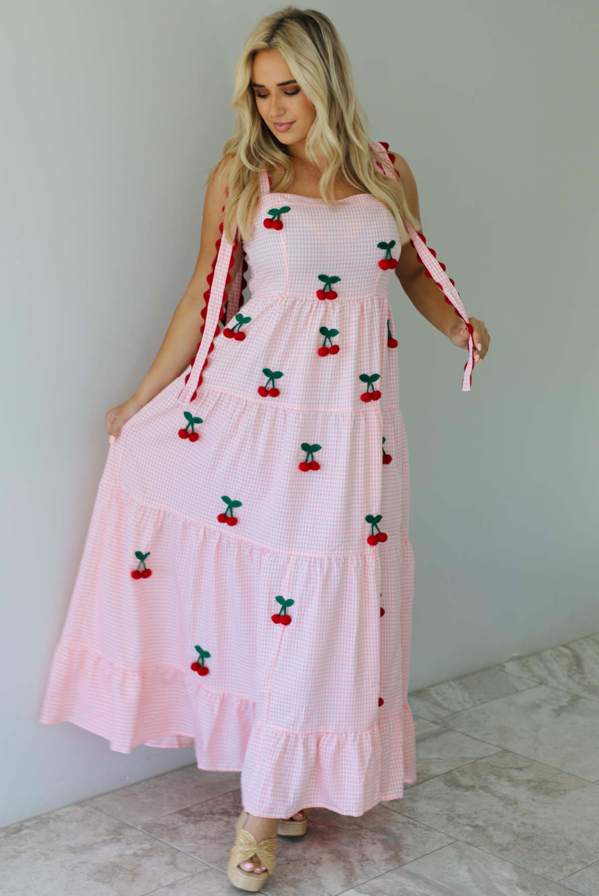 Woman wearing a pink and white gingham  maxi dress with thick straps, a fitted bodice, flowy skirt, and crocheted cherry details on the front indoors against a white wall with white marble flooring. 