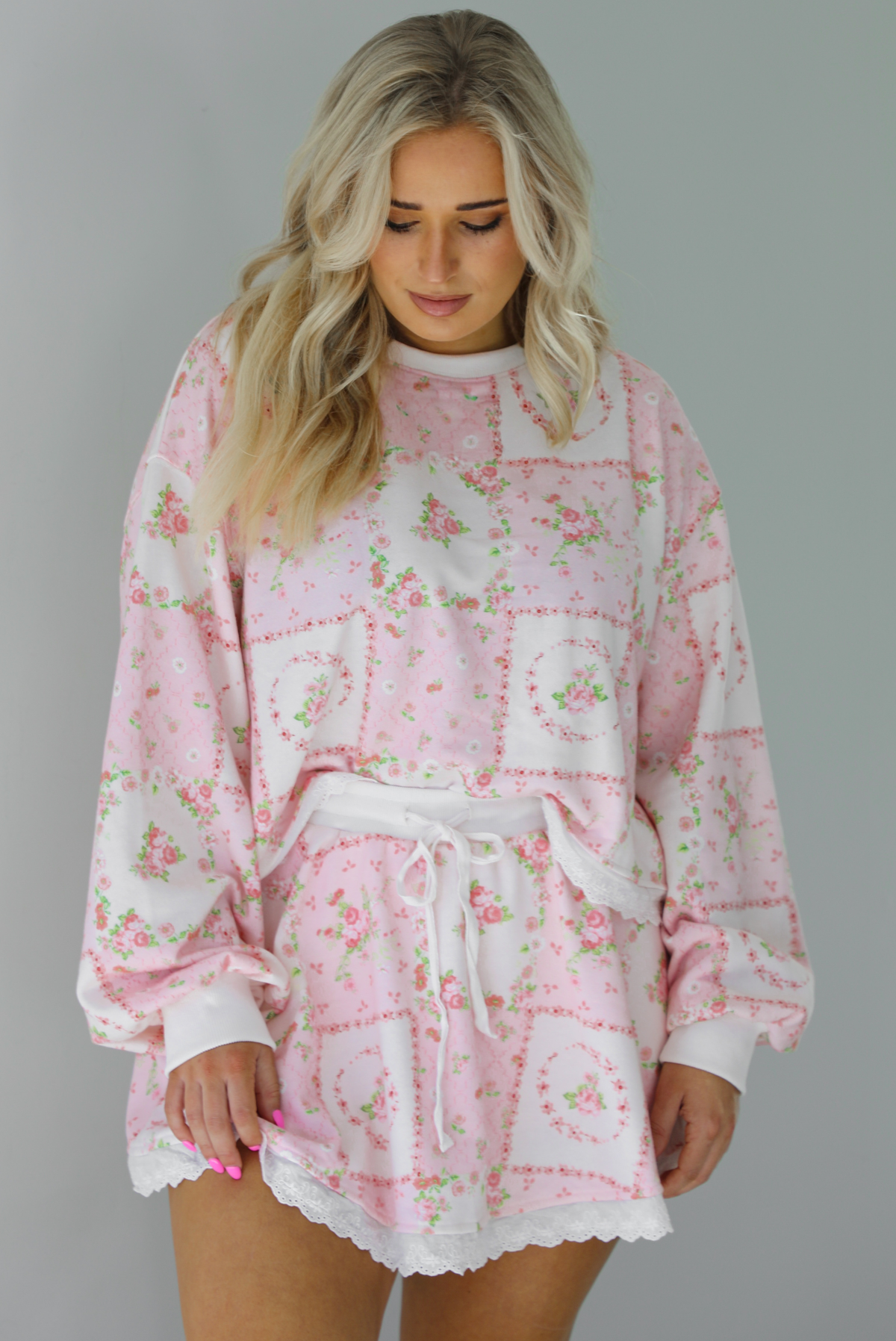Woman wearing a two piece set featuring a pink floral set that has an oversized long sleeved top and high waisted skirt. The photo is taken indoors against a white wall.