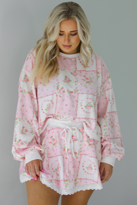 Woman wearing a two piece set featuring a pink floral set that has an oversized long sleeved top and high waisted skirt. The photo is taken indoors against a white wall.