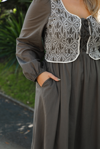 Close up of a woman model wearing an olive and off-white stitched detail dress outdoors. Dress has a corset-appearance bodice, long sleeves, functional pockets on either side, an elastic waistline, and a long, maxi length.