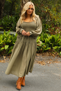 Woman model wearing an olive colored maxi length dress that has a smocked bodice, long sleeves that cinch with elastic at the wrists, and a flowy skirt. The photo is taken outdoors with greenery in the back.
