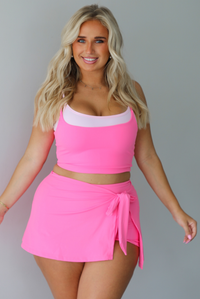A woman modeling a bright pink two-piece swimwear set consisting of a scoop-neck crop top with a light pink trim and a matching high-waisted wrap mini skirt with a side tie, standing indoors against a white wall.