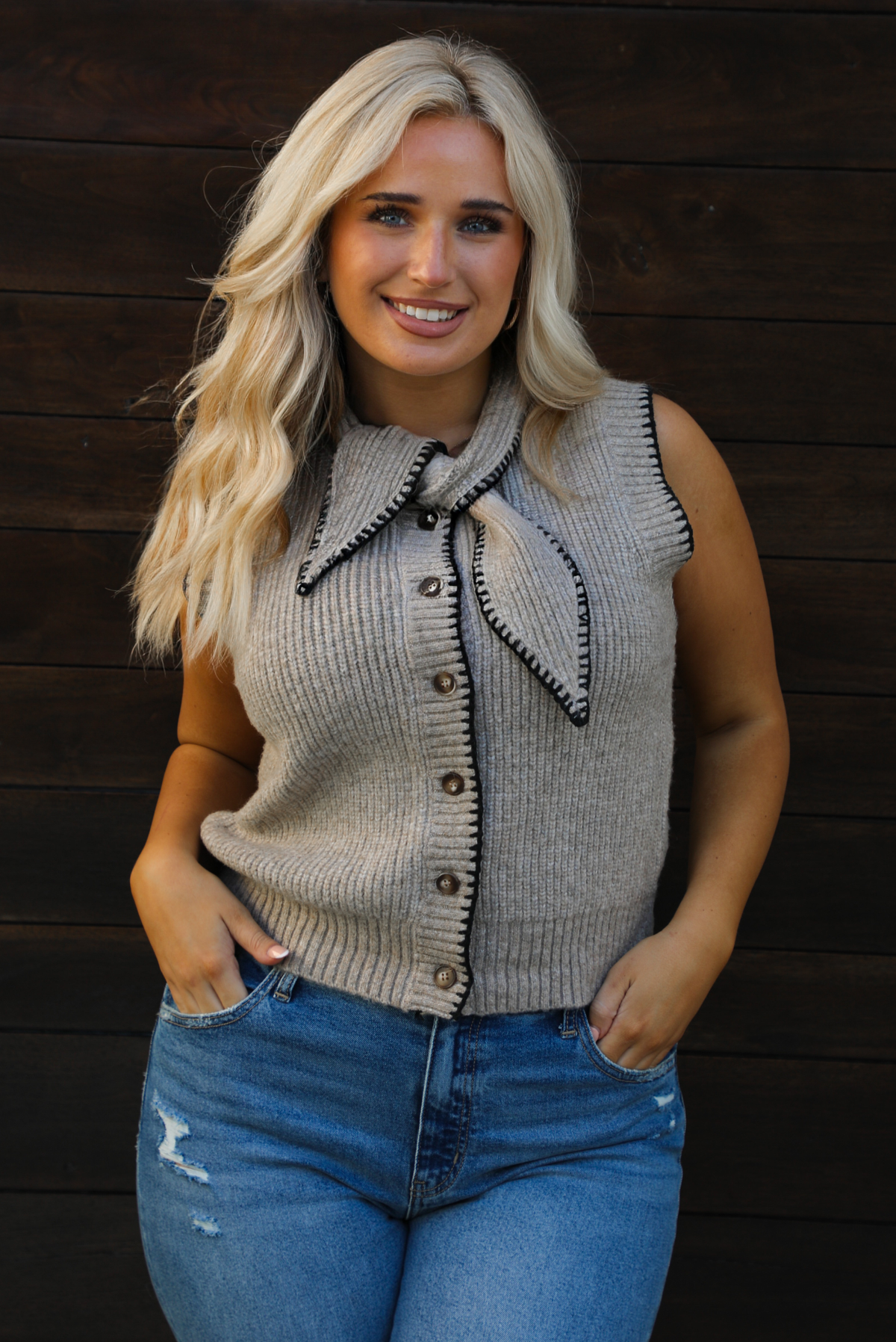 Close up of a woman model wearing a taupe sweater vest with black stitched detailing, has a tie detail at the neckline, is sleeveless, has functional button closures in the front, and has a ribbed texture. The sweater vest is paired with a medium wash denim. The photo is taken outdoors against a dark brown wooden door. 
