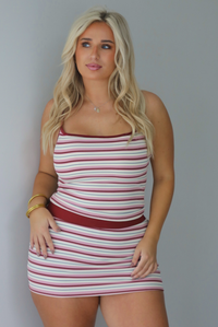 A woman wearing a berry, pink, white, and grey striped two piece set featuring a tank with a mini skirt, standing indoors against a white wall. 