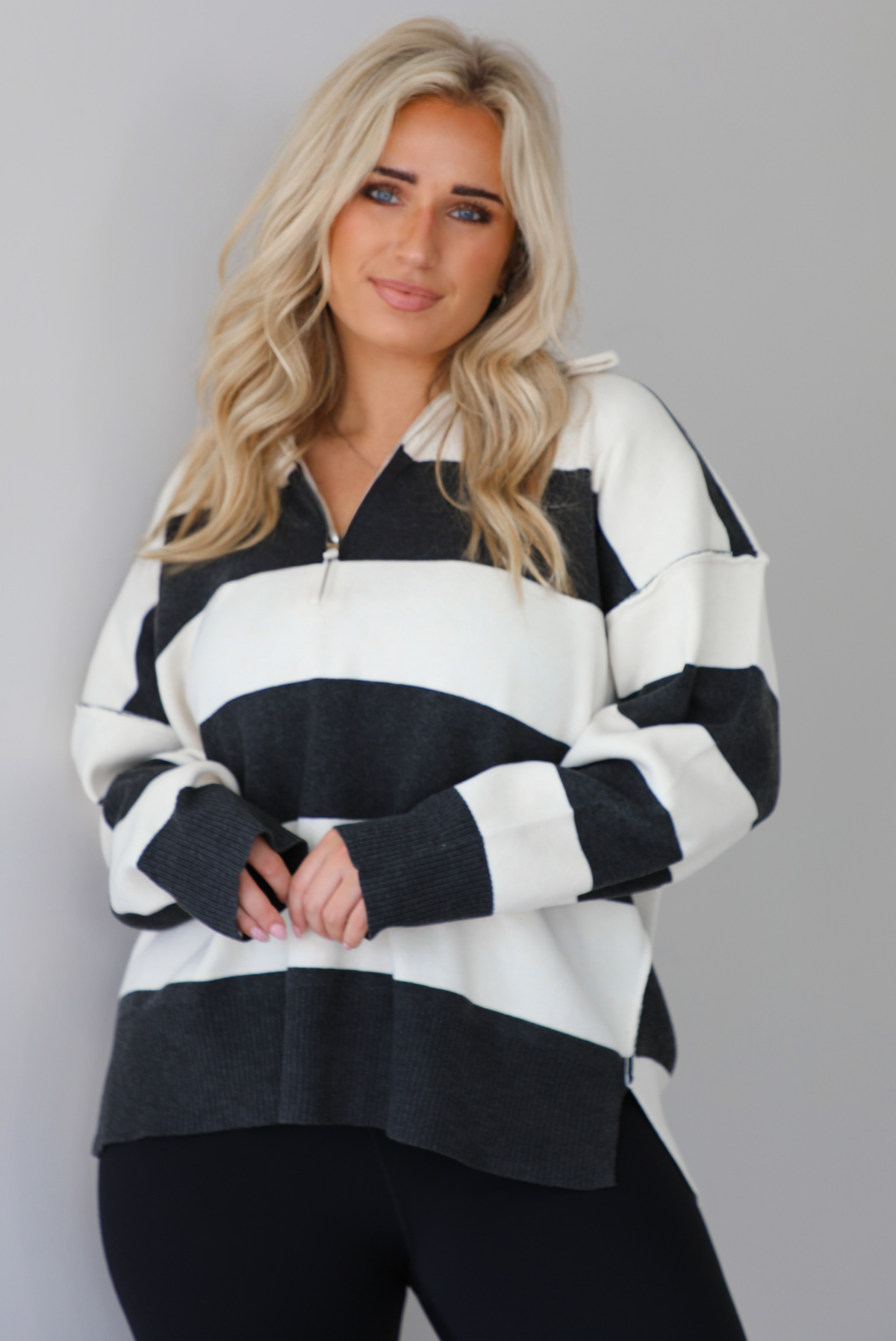 Woman wearing a charcoal and white striped quarter-zip sweater with a collared neckline, raw seam details, and a relaxed fit. It's paired with a fitted black jumpsuit. The photo is taken indoors against a white wall with white marble flooring.