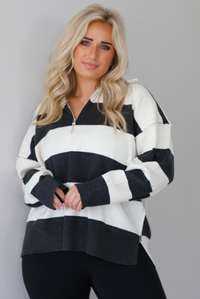 Woman wearing a charcoal and white striped quarter-zip sweater with a collared neckline, raw seam details, and a relaxed fit. It's paired with a fitted black jumpsuit. The photo is taken indoors against a white wall with white marble flooring.