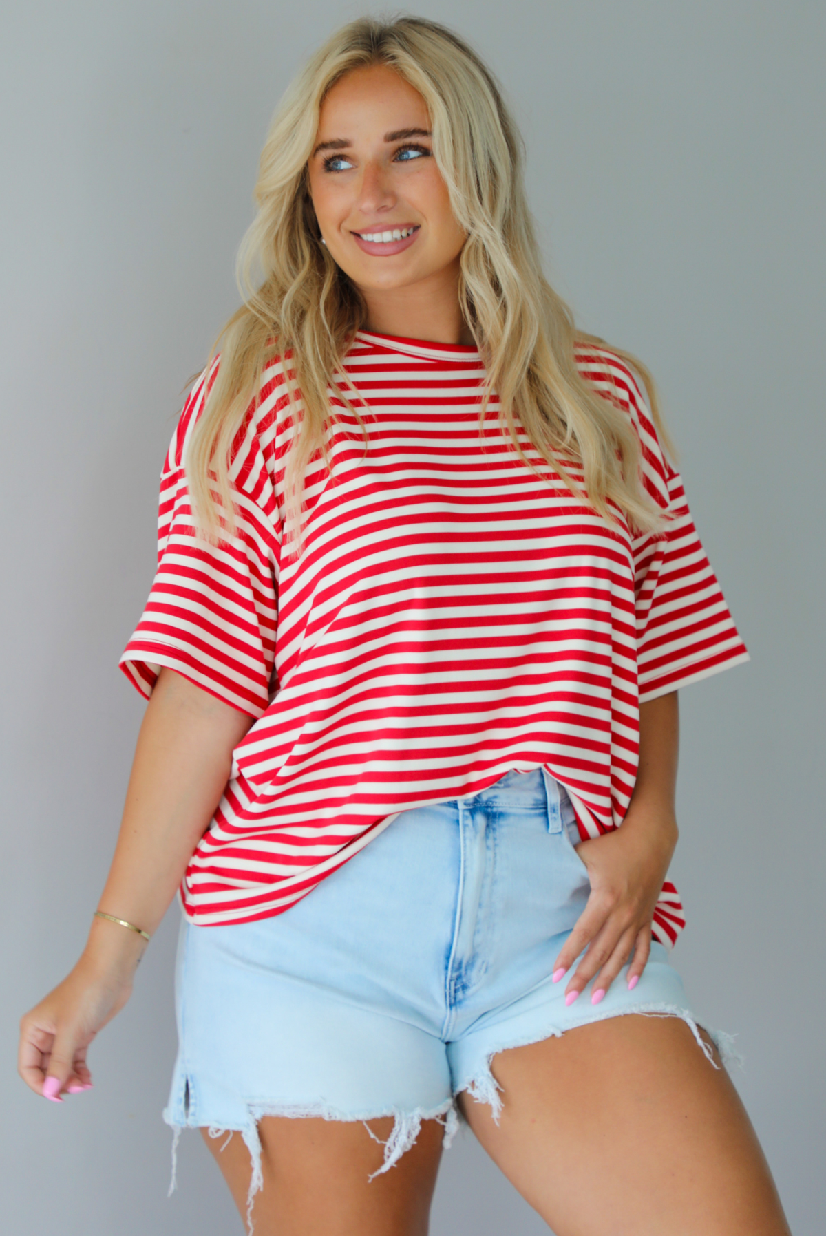 A woman modeling a red and white horizontally striped short-sleeve t-shirt with light blue denim shorts and brown sandals, standing indoors against a white wall. The top is tucked into the front of the shorts. 