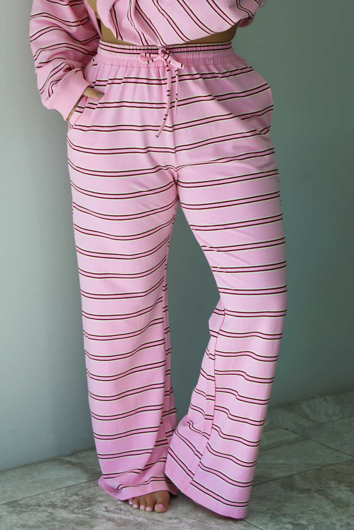 A woman wearing a pink two piece sweat set featuring a maroon and white striped design on a sweatshirt and high waisted, wide leg pants, standing indoors against a white wall with white marble flooring. The photo shows a closeup of the pants, which have an elastic and drawstring waistline and functional pockets on either side. 