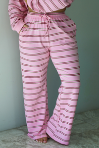 A woman wearing a pink two piece sweat set featuring a maroon and white striped design on a sweatshirt and high waisted, wide leg pants, standing indoors against a white wall with white marble flooring. The photo shows a closeup of the pants, which have an elastic and drawstring waistline and functional pockets on either side. 
