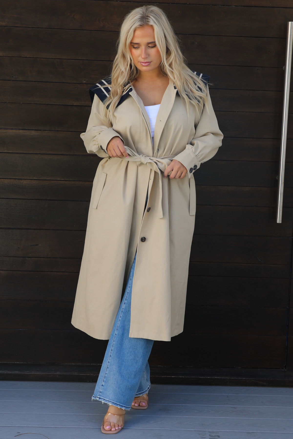 A woman wearing a tan trench coat with a navy collared neckline, functional button closures in the front, functional pockets on either side, with a tie waistline, paired with light wash flared hem denim jeans, standing outdoors in front of a dark wooden door. 