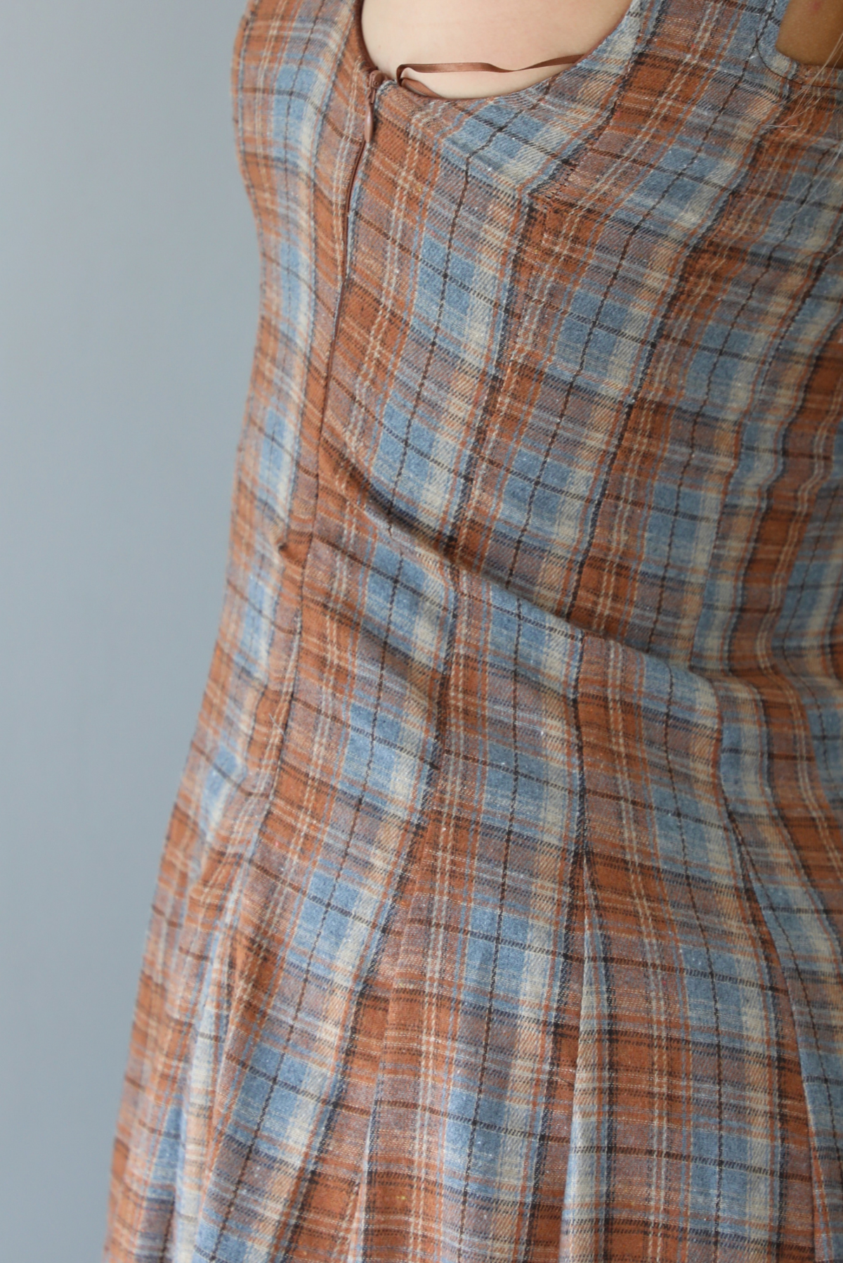 A woman wearing a brown and blue plaid midi dress with thick straps, a square neckline, fitted bodice that starts flaring out at the hips and ends about mid-shin, with a flowy skirt, standing indoors against a white wall. The photo shows a closeup of the zipper closure on the side. 