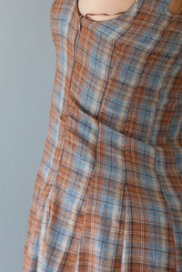 A woman wearing a brown and blue plaid midi dress with thick straps, a square neckline, fitted bodice that starts flaring out at the hips and ends about mid-shin, with a flowy skirt, standing indoors against a white wall. The photo shows a closeup of the zipper closure on the side. 