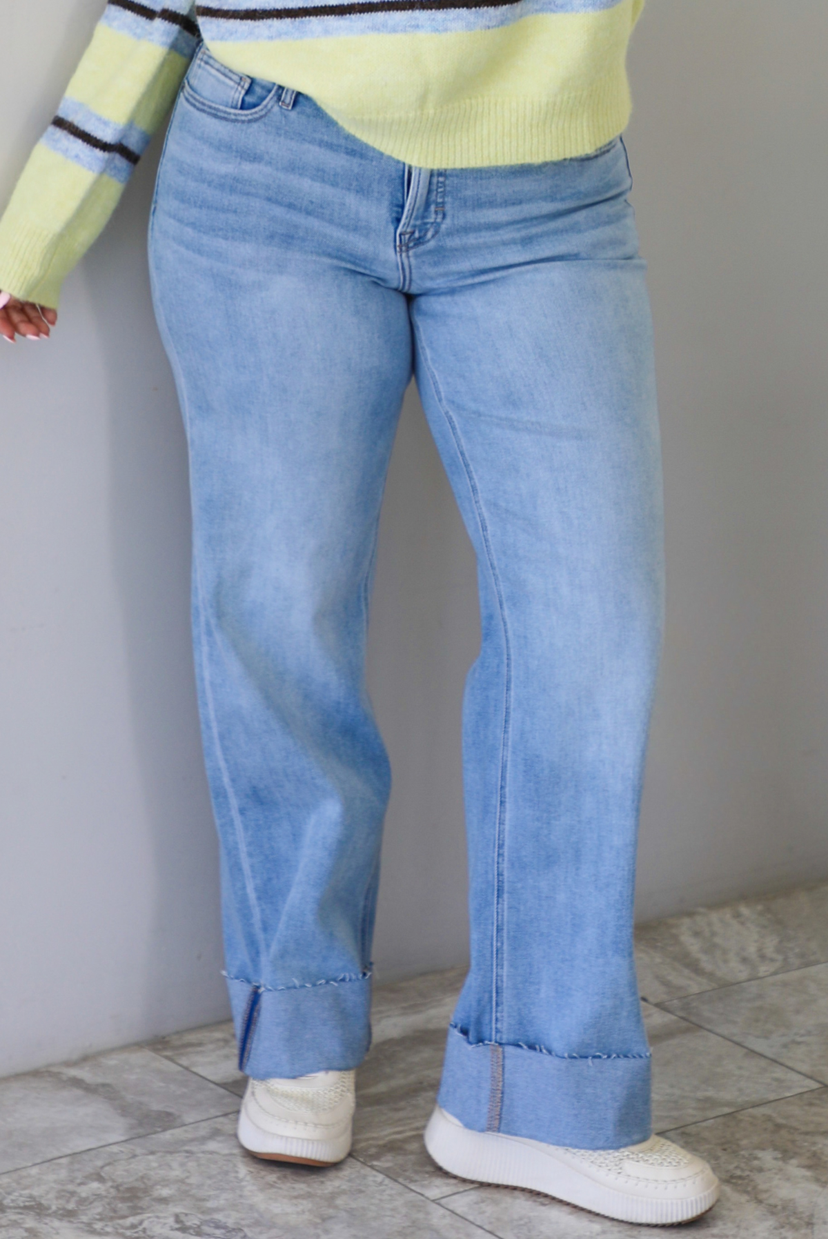 Woman wearing a pair of mid-rise light-wash denim that have a boot cut design and cuffed hem. The photo is taken indoors against a white wall with white marble flooring. 
