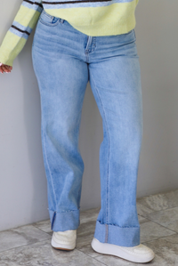 Woman wearing a pair of mid-rise light-wash denim that have a boot cut design and cuffed hem. The photo is taken indoors against a white wall with white marble flooring. 