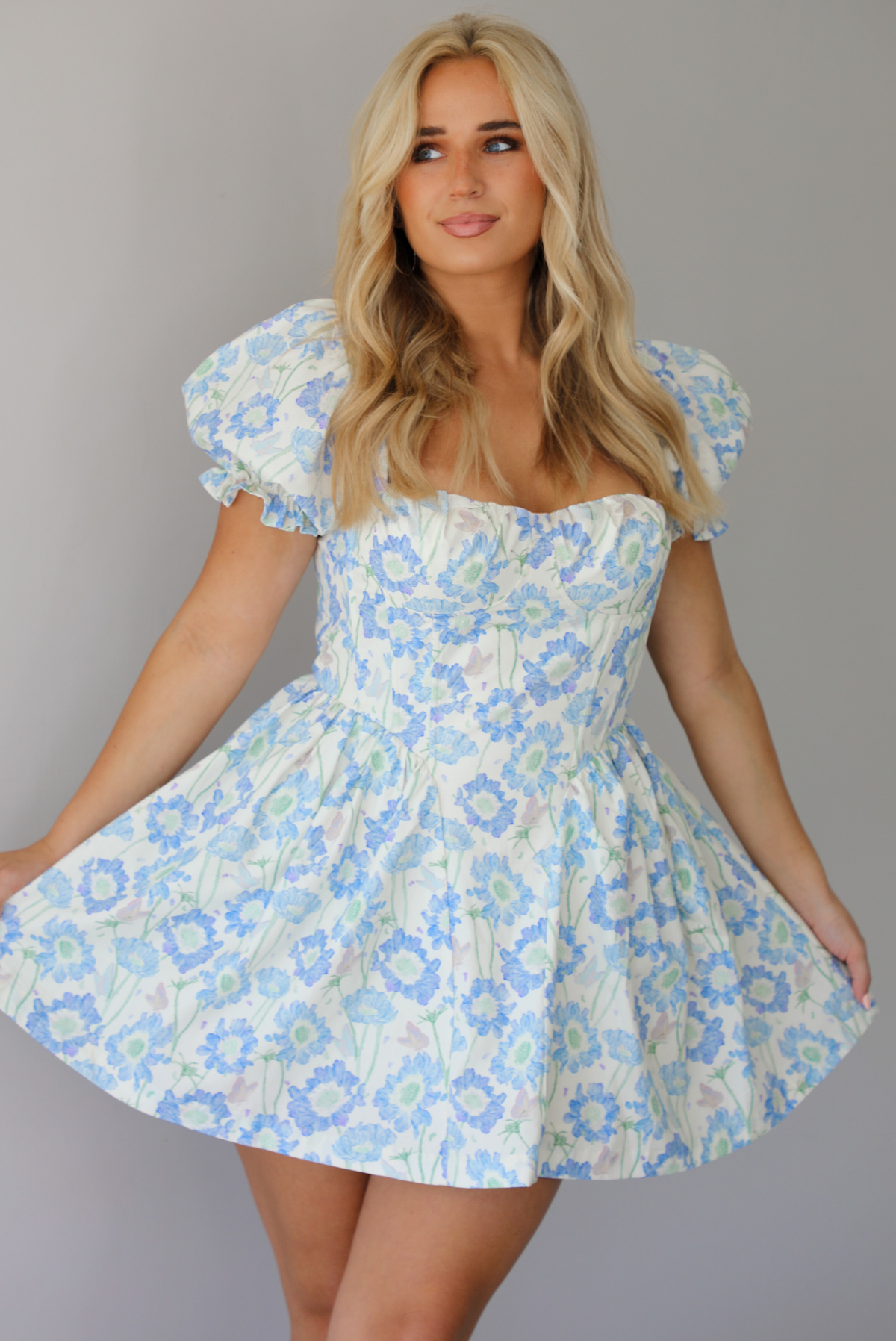 Woman wearing a white, blue, and green floral print mini dress that has short, puffed sleeves, a fitted corset bodice, and a flowy skirt indoors against a white wall.
