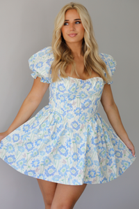 Woman wearing a white, blue, and green floral print mini dress that has short, puffed sleeves, a fitted corset bodice, and a flowy skirt indoors against a white wall.