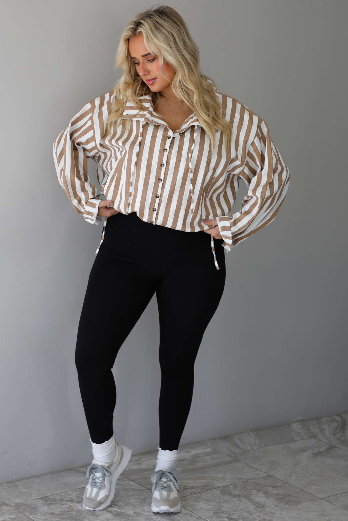 A woman wearing a tan and white striped collared long sleeved top with functional button closures along the front, worn with black leggings, white high ankle socks, and cream sneakers, standing indoors against a white wall with white marble flooring. 