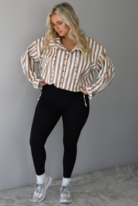 A woman wearing a tan and white striped collared long sleeved top with functional button closures along the front, worn with black leggings, white high ankle socks, and cream sneakers, standing indoors against a white wall with white marble flooring. 