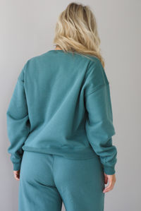 Woman wearing a sage two piece sweat-set. The set features a slightly oversized sweater and high waisted sweat pants that have an elastic and drawstring waistline with pockets. The sweater is untucked and shows the full length of the back of the sweater. The photo is taken indoors against a white wall with white marble flooring. 